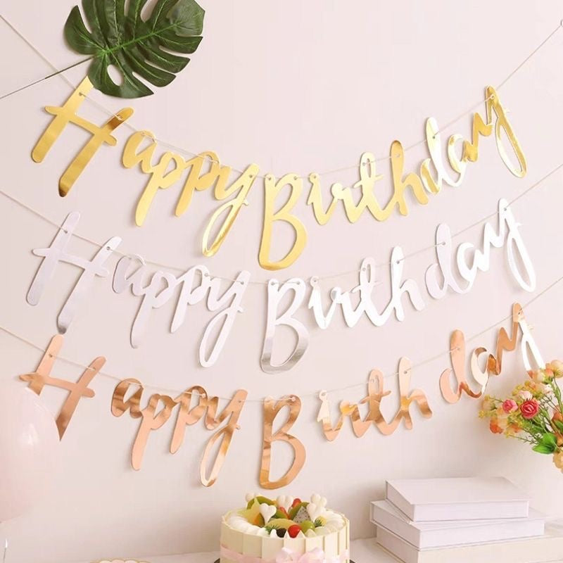 Gold 'Happy Birthday' letter decorations on a white wall with a cake and flowers.