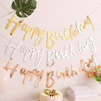 Gold 'Happy Birthday' letter decorations on a white wall with a cake and flowers.